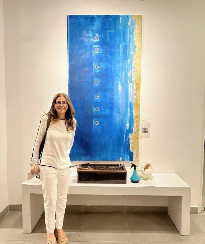 Elsa Basil standing beside her tall blue painting En el inmenso mar at an exhibition