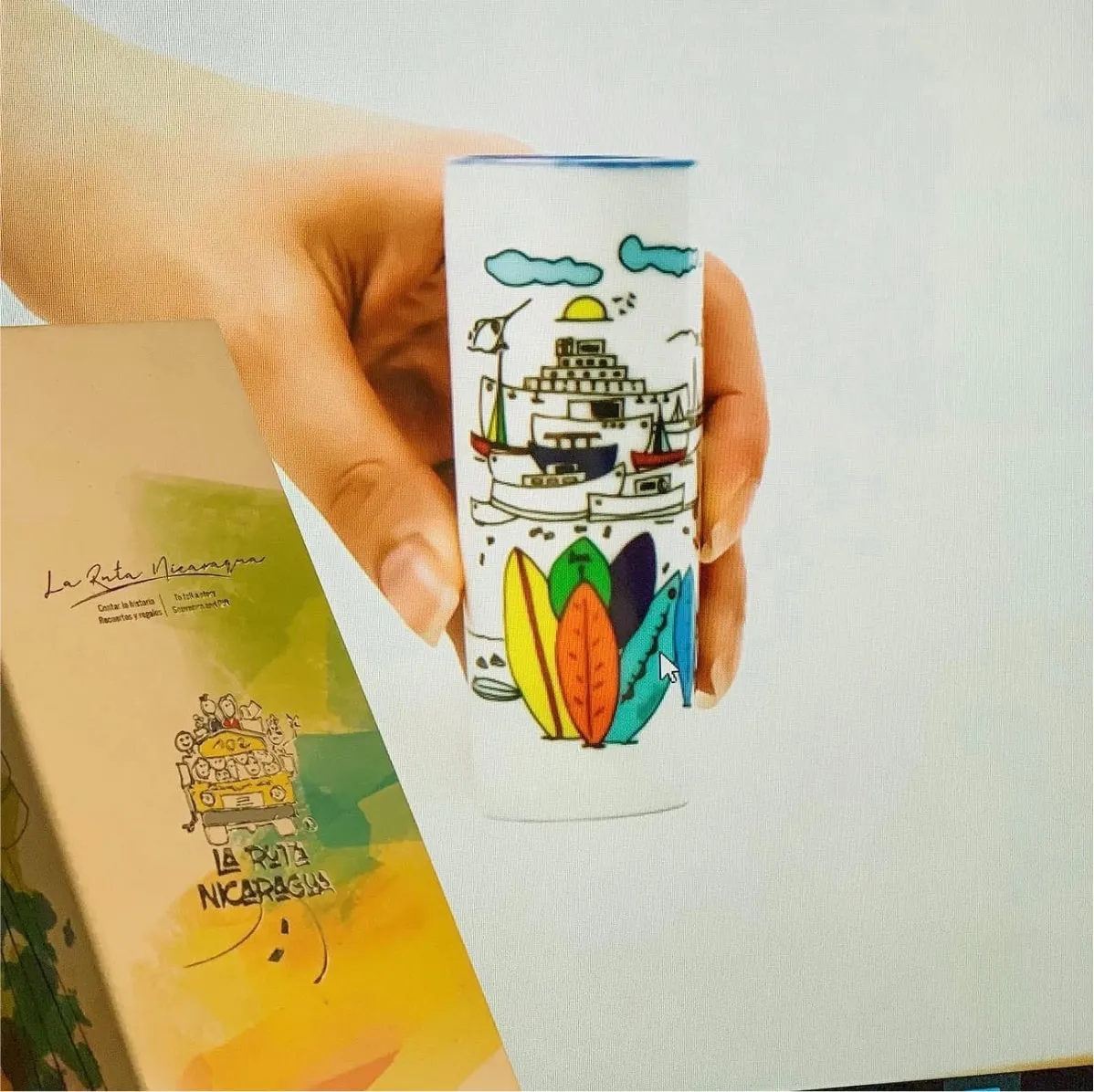 A hand holding a cup printed with the San Juan del Sur artwork, its illustrated card behind